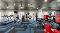 This fitness center is designed to support a comprehensive workout experience within a single, open-plan floor. The space is finished with a bold herringbone-patterned carpet in teal, navy, and charcoal tones, adding a dynamic visual energy to the room. The dark-painted ceiling, outfitted with recessed lighting and a ceiling fan, creates a sleek, focused atmosphere.

Natural light fills the space through multiple large windows along the walls, offering views of surrounding landscaped grounds. A full-length wall mirror on one side enhances the sense of openness and allows users to monitor their form during workouts.

The cardio zone features treadmills, an elliptical trainer, and a stationary bike, each positioned to take advantage of the outdoor views. A wall-mounted flat-screen television serves the cardio area for entertainment during workouts. An additional television is mounted on the opposite wall near the entry.

The strength training section includes a rack of dumbbells, a cable machine, and various resistance equipment. Exercise accessories such as medicine balls, stability balls, and a weight bench round out the offerings. A glass entry door is decorated with motivational fitness-themed typography, reinforcing the energetic tone of the space. A small refreshment and towel station provides added convenience for users.