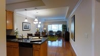 This single-floor residence showcases an open-concept design that seamlessly integrates the kitchen, dining, and living areas into one flowing space. The kitchen features dark granite countertops atop warm wood cabinetry, with a peninsula that acts as a natural divider between the cooking and dining zones. A chandelier with multiple lights hangs above the kitchen area, complementing a separate pendant light fixture, both adding warmth to the space.

Rich hardwood flooring extends throughout the entire layout, lending a sense of continuity and elegance. The dining area accommodates a full table with seating and sits adjacent to the kitchen, making it ideal for entertaining. Beyond the dining space, the living room is furnished with classic seating arrangements centered around a coffee table, anchored by a green area rug that adds a pop of color against the warm floors.

Large windows along the far wall of the living room allow generous natural light to fill the space, creating a bright and inviting atmosphere. Crown molding details along the ceiling and large-scale artwork on the walls add refined finishing touches, contributing to the overall polished and comfortable ambiance of the home.