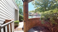 This single-floor residence offers a welcoming exterior defined by classic architectural details and a lush, well-maintained setting. The unit is clad in traditional lap siding in a soft gray-green tone, complemented by crisp white trim and a six-panel entry door that adds a timeless charm to the facade.

A standout feature of this space is the covered patio area, framed by a substantial full brick column and a curved decorative brick half-wall with a lattice-style cutout pattern. This thoughtfully designed outdoor area provides both privacy and an open connection to the surrounding landscape, with mature trees and manicured shrubs creating a natural, tranquil atmosphere.

The patio extends the living space outdoors and is accessible via a sliding glass door screened for ventilation, making it ideal for relaxing while enjoying the outdoor setting. From this vantage point, residents are treated to direct views of a sparkling community pool and a covered clubhouse, enhancing the sense of resort-style living.

The grounds surrounding the unit feature river rock accents, decorative mulched beds, and a mix of ornamental shrubs that lend a polished, curated look to the exterior. A secondary entry door with a painted wood railing and concrete step provides an additional access point. The overall ambiance is peaceful and inviting, blending classic brick detailing with lush greenery and community amenities just steps away.