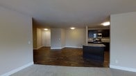 This single-floor residence offers an open-concept layout that connects the living and kitchen areas in a seamless, functional design. The living space is finished with rich, dark hardwood flooring that adds warmth and a sense of depth to the room, while light-colored carpeting defines the entry transition zone, creating a natural boundary between spaces.

The kitchen is well-appointed with dark cabinetry that provides a striking contrast against the neutral walls and light countertops. Stainless steel appliances, including a range and over-the-range microwave, lend a modern touch, while a dark kitchen island serves as a central workspace and a visual anchor for the open layout. The cabinetry provides ample storage, and the overall configuration supports both everyday cooking and entertaining.

The living area is generously proportioned, offering flexibility for a variety of furniture arrangements. Neutral beige walls and flush-mount ceiling lighting contribute to a clean, understated aesthetic throughout. A textured ceiling adds subtle visual interest overhead.

Toward the back of the space, a hallway leads to additional rooms, including what appears to be a closet or utility area with classic paneled doors. The overall ambiance is practical and inviting, combining durable finishes with a cohesive, contemporary design sensibility.