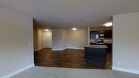 This single-floor residence offers an open-concept layout that connects the living and kitchen areas in a seamless, functional design. The living space is finished with rich, dark hardwood flooring that adds warmth and a sense of depth to the room, while light-colored carpeting defines the entry transition zone, creating a natural boundary between spaces.

The kitchen is well-appointed with dark cabinetry that provides a striking contrast against the neutral walls and light countertops. Stainless steel appliances, including a range and over-the-range microwave, lend a modern touch, while a dark kitchen island serves as a central workspace and a visual anchor for the open layout. The cabinetry provides ample storage, and the overall configuration supports both everyday cooking and entertaining.

The living area is generously proportioned, offering flexibility for a variety of furniture arrangements. Neutral beige walls and flush-mount ceiling lighting contribute to a clean, understated aesthetic throughout. A textured ceiling adds subtle visual interest overhead.

Toward the back of the space, a hallway leads to additional rooms, including what appears to be a closet or utility area with classic paneled doors. The overall ambiance is practical and inviting, combining durable finishes with a cohesive, contemporary design sensibility.