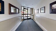 This single-floor corridor space functions as a transitional lobby and hallway system, thoughtfully designed to guide occupants through the building with ease and clarity. The layout follows an L-shaped or cross configuration, connecting an exterior entrance vestibule to interior hallway wings that lead deeper into the building.

The entrance vestibule is anchored by a set of large glass doors with aluminum framing, allowing natural light to pour in and offering views of the surrounding outdoor greenery. A durable carpet runner leads visitors from the entry threshold inward, complementing the light-toned terrazzo flooring found in adjacent sections.

The hallways are finished with two-tone walls — a warm beige lower half divided by a white chair rail, and a lighter upper half — creating a clean, professional aesthetic. Recessed and flush-mounted ceiling lights provide consistent, even illumination throughout. Framed artwork and mirrors are thoughtfully placed along the corridor walls, adding a welcoming character to the space.

One section of the corridor features distinctive green mosaic tile walls, lending a contrasting architectural texture that differentiates this wing visually. Safety features including fire alarm pull stations and a wall-mounted hand sanitizer dispenser are also present. Interior doors with glazed panels provide access to additional areas of the building, maintaining visual connectivity while defining clear transitions between zones.
