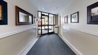This single-floor corridor space functions as a transitional lobby and hallway system, thoughtfully designed to guide occupants through the building with ease and clarity. The layout follows an L-shaped or cross configuration, connecting an exterior entrance vestibule to interior hallway wings that lead deeper into the building.

The entrance vestibule is anchored by a set of large glass doors with aluminum framing, allowing natural light to pour in and offering views of the surrounding outdoor greenery. A durable carpet runner leads visitors from the entry threshold inward, complementing the light-toned terrazzo flooring found in adjacent sections.

The hallways are finished with two-tone walls — a warm beige lower half divided by a white chair rail, and a lighter upper half — creating a clean, professional aesthetic. Recessed and flush-mounted ceiling lights provide consistent, even illumination throughout. Framed artwork and mirrors are thoughtfully placed along the corridor walls, adding a welcoming character to the space.

One section of the corridor features distinctive green mosaic tile walls, lending a contrasting architectural texture that differentiates this wing visually. Safety features including fire alarm pull stations and a wall-mounted hand sanitizer dispenser are also present. Interior doors with glazed panels provide access to additional areas of the building, maintaining visual connectivity while defining clear transitions between zones.