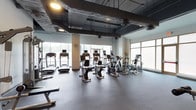 This fitness center offers a spacious, single-floor layout designed to accommodate a comprehensive cardio workout experience. The facility is outfitted with a robust selection of exercise equipment, including multiple treadmills, stationary bikes, elliptical trainers, and stair climbers, all arranged to allow comfortable movement between machines. The flooring consists of durable rubber matting, well-suited for a high-traffic fitness environment. Overhead, an industrial-style exposed ceiling reveals ductwork, structural elements, and recessed lighting fixtures that provide consistent illumination throughout the space. Large floor-to-ceiling windows line the perimeter walls, inviting abundant natural light and contributing to an open, airy atmosphere. A support column near the center of the room adds a structural architectural element without impeding the overall flow of the space. The combination of modern equipment, generous natural light, and a practical industrial aesthetic makes this fitness center a functional and well-appointed facility suitable for a variety of cardio training routines.