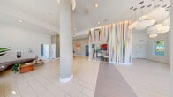 Located at 2500 SW 22nd Ter in Fort Lauderdale, Florida, this single-floor space is a striking example of contemporary design that seamlessly blends artistry with functionality. The interior is immediately distinguished by its soaring double-height ceilings, dramatic floor-to-ceiling windows, and circular porthole accents that flood the space with an abundance of natural light and frame lush views of surrounding palm trees and tropical landscaping.

The main living and lounge area serves as the heart of the space, featuring light-toned large-format tile flooring, a curated mix of modern seating including swivel accent chairs and a curved lounge sofa, and a bold, multicolored vertical striped wall mural that adds a dynamic artistic focal point. Globe pendant chandeliers and wall-mounted red sconces provide layered, stylish lighting throughout.

A glass-partitioned office area offers a dedicated workspace with a refined, professional atmosphere, while maintaining visual connectivity to the main floor through its transparent walls. The hallway provides a clean transitional corridor connecting the primary areas to the ADA-accessible bathroom, which is finished with large-format dark tile flooring and walls, a wall-mounted sink, and a clean, utilitarian aesthetic.

Overall, the space exudes a bright, modern, and energetic ambiance — an inviting environment defined by artistic expression, ample natural light, and thoughtful design.