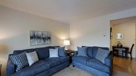 This single-story space presents a well-appointed living area designed for comfort and functionality. The living room is furnished with two matching navy blue sofas, each dressed with an assortment of decorative pillows in complementary patterns, including geometric and paisley designs, creating a cohesive and inviting aesthetic. A small dark-finished side table between the sofas holds a classic table lamp, providing warm ambient lighting that enhances the room's relaxed atmosphere. The walls are painted in a neutral, soft tone that pairs seamlessly with the carpeted flooring, contributing to an overall sense of calm. A framed abstract artwork in muted tones of black, white, and blue anchors one wall, adding a subtle artistic element to the space. The ceiling is smooth and standard in height, maintaining an open feel throughout. A wall-mounted control panel, likely for climate or smart home features, is also visible, indicating modern convenience. Overall, the living area is thoughtfully arranged to maximize comfort while maintaining a clean and organized appearance.
