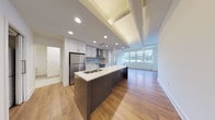 Located at 1240 Huron Rd E in Cleveland, Ohio, this single-floor unit offers a thoughtfully designed open-concept layout that blends modern finishes with functional living spaces. The kitchen serves as a central focal point, featuring a large island with a white quartz countertop, dark base cabinetry, white upper cabinets, stainless steel appliances including a refrigerator and range, a tile backsplash, and a stainless steel wall-mounted range hood. Recessed lighting illuminates the space throughout, while exposed ductwork along the ceiling adds an industrial character. The open living area extends from the kitchen, bathed in natural light from large windows that frame views of the surrounding Cleveland cityscape. Warm hardwood-style flooring runs consistently through the main living areas, contributing to a cohesive aesthetic. The bedroom features its own window with natural light and hardwood-style flooring. A spacious walk-in closet offers wire shelving and carpeted flooring for comfortable storage. The bathroom includes light tile flooring, a vanity with built-in storage, and a tub/shower combination. An in-unit washer and dryer are also present, adding everyday convenience to this well-appointed urban residence.