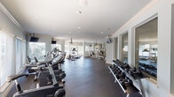 This fitness center in Spring, TX offers a comprehensive workout environment designed to accommodate a wide range of exercise needs. The space is thoughtfully organized into distinct functional zones, all sharing a single open floor plan finished with durable dark rubber flooring throughout.

The cardio zone is generously stocked with treadmills, elliptical trainers, stationary bikes, and rowing machines, arranged to maximize spacing between equipment. Large windows with blinds line multiple walls, allowing substantial natural light to fill the space while providing views of the surrounding community. Ceiling-mounted fans are positioned throughout, aiding air circulation during workouts.

A dedicated strength training section features an extensive selection of selectorized cable and resistance machines arranged in parallel rows, complemented by a full dumbbell rack offering a wide weight range. Large wall mirrors visually expand this area and allow users to monitor their form during exercises.

Multiple flat-screen televisions are mounted throughout the facility, both on walls and from ceiling brackets, keeping users entertained during workouts. Crown molding and soft blue-gray walls lend a polished, refined aesthetic that distinguishes this fitness center from more utilitarian gym environments. Recessed lighting supplements the natural light, ensuring the space remains well-illuminated at all times.