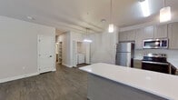 This single-story, 2-bedroom, 2-bathroom residence offers a thoughtfully designed layout with modern finishes and functional living spaces. The open-concept kitchen and living area serves as the heart of the home, featuring dark hardwood-style flooring, taupe cabinetry, stainless steel appliances including a refrigerator and dishwasher, and a sleek white countertop island with pendant lighting overhead. The living room extends naturally from the kitchen, bathed in natural light from large windows with blinds, and is equipped with a ceiling fan for added comfort. A glass exterior door provides convenient outdoor access directly from the living area. The spacious primary bedroom is carpeted and generously sized, accompanied by a walk-in closet with built-in hanging rods. The second bedroom also features carpet and a large window that fills the room with natural light. Both bathrooms are finished with taupe cabinetry vanities and wood-style tile flooring. A dedicated in-unit laundry area accommodates a full-sized washer and dryer side by side. The hallway connects all rooms efficiently, and two closets provide ample storage throughout. The neutral color palette and consistent finishes lend the space a clean, cohesive aesthetic.