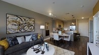 This single-floor space showcases a thoughtfully designed open-concept layout that connects the living, dining, and kitchen areas into one cohesive environment. The living area features warm hardwood-style flooring, neutral gray walls, and is anchored by a plush dark sofa accented with decorative pillows in muted tones and pops of gold. A round white coffee table serves as a central focal point, complemented by a textured area rug beneath. Bold abstract wall art adds a striking visual element to the space.

The dining area transitions naturally from the living space, featuring a round white dining table surrounded by upholstered chairs, creating an intimate yet functional setting. Pendant lighting overhead adds warmth and definition to the zone.

The kitchen is outfitted with light wood cabinetry, stainless steel appliances including a refrigerator and microwave, and a light-toned countertop with bar seating. Track lighting runs along the ceiling, providing even illumination throughout. The overall design palette of warm neutrals, wood tones, and modern fixtures creates a cohesive and contemporary ambiance.