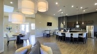 This single-floor space is designed as a contemporary open-concept lounge and social hub. The layout flows seamlessly from a comfortable seating area in the foreground — anchored by a plush gray sofa accented with yellow throw pillows — to a series of distinct gathering zones that extend throughout the room.

Large drum pendant lights hang prominently from the soaring ceiling, casting warm, even illumination across the hardwood-style flooring below. Recessed lighting and additional pendant fixtures further enhance the bright, welcoming atmosphere. Neutral gray walls are complemented by a bold dark accent wall near the bar area, adding visual depth and contrast.

A long bar-height counter with white upholstered stools serves as a central feature, backed by dark cabinetry and countertop appliances. Multiple high-top and standard dining tables with upholstered chairs are distributed throughout the space, offering flexible seating arrangements.

A wall-mounted flat-screen TV is positioned for easy viewing from multiple vantage points. Natural light streams in through glass-paned doors along one wall, connecting the interior to an outdoor area beyond. The overall ambiance is polished, modern, and functional.