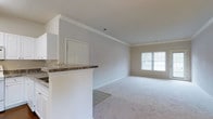 This single-floor space offers a clean, neutral aesthetic defined by light gray walls, plush wall-to-wall carpeting, and white trim throughout. Crown molding lines the ceilings, adding a refined architectural detail to each room and contributing to an overall sense of polish.

The open-concept living area is bright and airy, benefiting from a large window that allows natural light to fill the space. Adjoining the living area is a kitchen featuring white cabinetry, a raised peninsula with a granite-style countertop, and tile flooring, creating a natural transition between the kitchen and the carpeted living space. The peninsula provides both a functional workspace and a casual dining or serving area.

A hallway extends from the main living space, lined with white doors and illuminated by ceiling-mounted light fixtures. The corridor connects to additional rooms, including what appears to be a closeted space with louvered doors — ideal for storage. The consistent color palette of soft grays and whites throughout the space creates a cohesive, move-in-ready environment suited to a variety of interior styles.