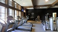 This fitness center combines a classic architectural aesthetic with a comprehensive selection of modern exercise equipment. The space is defined by its striking exposed dark wood beam ceiling, which adds warmth and character, while brick accent walls lend an industrial charm that sets it apart from a typical gym environment. Large arched and floor-to-ceiling windows line the walls, inviting abundant natural light and offering views of the surrounding greenery, creating an energizing atmosphere for workouts.

The cardio zone is well-appointed with multiple Cybex treadmills and elliptical machines arranged to maximize space and airflow. A mounted flat-screen television is positioned for easy viewing during cardio sessions. Strength training is supported by a dedicated cable and weight machine station, a free weight bench, and a rowing machine, providing a well-rounded fitness experience.

A bold black accent wall serves as a focal point, adorned with an athletic mural that reinforces the motivational spirit of the space. Recessed lighting supplements the natural light, ensuring the room remains bright and functional at any hour. The durable fitness flooring throughout provides safety and comfort during all types of exercise. Overall, this fitness center delivers a distinctive, inviting environment that encourages an active lifestyle.