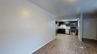 This single-floor space offers an open-concept layout that seamlessly connects the living area to the kitchen, creating a sense of spaciousness and functionality. The flooring throughout is a light wood-tone laminate or vinyl plank, providing a clean, contemporary aesthetic that ties the space together cohesively.

The kitchen is a standout feature, outfitted with a full suite of stainless steel appliances, including a refrigerator, microwave, dishwasher, and range. Dark espresso-toned cabinetry lines the walls, offering ample storage while providing a striking contrast against the light-colored walls and flooring. A sink is positioned along the counter, and the layout allows for efficient workflow within the kitchen zone.

The open living area is defined by smooth, light gray walls and white baseboards, giving the space a clean and neutral palette that lends itself well to a variety of furnishing styles. Recessed lighting illuminates the kitchen area, while the overall interior maintains a bright, well-lit atmosphere. The simple, modern design of this space makes it highly adaptable and move-in ready.