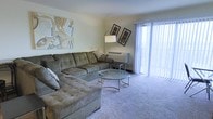 This single-floor residence offers a welcoming living room as its central gathering space. The room is finished with soft, neutral-toned carpeting that adds warmth underfoot and complements the light-colored walls. A large L-shaped sectional sofa in a muted olive-gray upholstery serves as the focal point, providing generous seating and a relaxed atmosphere. Centered before the sofa sits a round glass-top coffee table with a decorative metal frame, adding a subtle contemporary touch to the room.

Natural light is a notable feature, courtesy of floor-to-ceiling sliding glass doors dressed with vertical blinds, which offer both privacy and access to the outdoors. The doors allow daylight to filter into the space, brightening the overall interior. A wall-mounted HVAC unit beneath the windows provides climate control. Decorative wall art, including an abstract canvas and two framed prints, adds personality to the neutral walls. A freestanding arc floor lamp contributes additional ambient lighting. Overall, the living area presents a comfortable and functional layout suitable for everyday living.