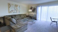 This single-floor residence offers a welcoming living room as its central gathering space. The room is finished with soft, neutral-toned carpeting that adds warmth underfoot and complements the light-colored walls. A large L-shaped sectional sofa in a muted olive-gray upholstery serves as the focal point, providing generous seating and a relaxed atmosphere. Centered before the sofa sits a round glass-top coffee table with a decorative metal frame, adding a subtle contemporary touch to the room.

Natural light is a notable feature, courtesy of floor-to-ceiling sliding glass doors dressed with vertical blinds, which offer both privacy and access to the outdoors. The doors allow daylight to filter into the space, brightening the overall interior. A wall-mounted HVAC unit beneath the windows provides climate control. Decorative wall art, including an abstract canvas and two framed prints, adds personality to the neutral walls. A freestanding arc floor lamp contributes additional ambient lighting. Overall, the living area presents a comfortable and functional layout suitable for everyday living.