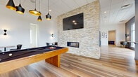 This contemporary amenity space is thoughtfully designed across a single floor, offering a versatile and inviting environment for both recreation and collaboration. The open layout is anchored by a striking stacked-stone accent wall that stretches to the ceiling, housing a linear fireplace at its base and a large flat-screen television above — making it the visual centerpiece of the room.

Wide-plank wood-look flooring runs throughout, lending warmth and cohesion to the space. A full-size pool table with a solid wood base occupies the recreational zone, complemented by stylish pendant lighting overhead with matte black and gold-interior fixtures that add a modern edge.

A long conference-style dining table with upholstered chairs provides a functional area for meetings or group dining, also bathed in coordinating pendant lighting. Along one wall, a built-in banquette with teal accent pillows and small bistro tables creates a relaxed lounge nook, adorned with a trio of framed artwork above.

Floor-to-ceiling glass doors open to an outdoor terrace, flooding the interior with natural light and offering views of the surrounding landscape. A utility area with countertops and small appliances provides added convenience. The overall ambiance is clean, modern, and welcoming.