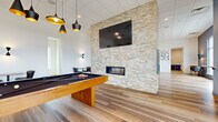 This contemporary amenity space is thoughtfully designed across a single floor, offering a versatile and inviting environment for both recreation and collaboration. The open layout is anchored by a striking stacked-stone accent wall that stretches to the ceiling, housing a linear fireplace at its base and a large flat-screen television above — making it the visual centerpiece of the room.

Wide-plank wood-look flooring runs throughout, lending warmth and cohesion to the space. A full-size pool table with a solid wood base occupies the recreational zone, complemented by stylish pendant lighting overhead with matte black and gold-interior fixtures that add a modern edge.

A long conference-style dining table with upholstered chairs provides a functional area for meetings or group dining, also bathed in coordinating pendant lighting. Along one wall, a built-in banquette with teal accent pillows and small bistro tables creates a relaxed lounge nook, adorned with a trio of framed artwork above.

Floor-to-ceiling glass doors open to an outdoor terrace, flooding the interior with natural light and offering views of the surrounding landscape. A utility area with countertops and small appliances provides added convenience. The overall ambiance is clean, modern, and welcoming.