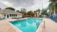 This outdoor pool area presents a generously sized swimming pool as its central feature, filled with clear, turquoise water that reflects the surrounding environment. The pool is well-defined with tiled edges and includes stainless steel handrails for easy entry and exit, with depth markers visible along the sides. The surrounding deck is finished in light-toned pavers, providing ample space for lounging and circulation. A row of dark-framed chaise lounges lines the perimeter, offering dedicated relaxation areas for guests. A small seating arrangement with teal chairs and a shaded umbrella table is positioned to one side, adding a casual social zone. The area is enclosed by a metal security fence, ensuring safety and privacy. Tall, mature palm trees and lush tropical greenery frame the entire space, lending a resort-like ambiance characteristic of a warm, southern climate. A low-profile building with large windows is situated adjacent to the pool deck, likely serving as a facility or common area. The overall atmosphere is open, airy, and tropical, making this pool area a well-appointed outdoor amenity suited for leisure and recreation.
