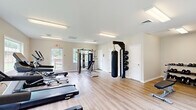 Located at 16 Wedgewood Dr in Spring Lake, NC, this single-floor community amenity building houses two distinct functional spaces: a fitness center and a laundry facility. The fitness center is bright and welcoming, featuring light hardwood-style flooring and large windows that invite abundant natural light and views of the surrounding greenery. The workout area is well-appointed with a range of equipment, including Spirit-brand treadmills, an elliptical machine, a multi-function cable/weight machine, an adjustable bench, a heavy punching bag with wall-mounted medicine ball rack, a dumbbell rack, and a rowing machine. Overhead fluorescent lighting ensures the space is well-lit throughout the day and evening. The laundry room offers a clean, functional environment finished with neutral tile flooring and a light, airy color palette. It is equipped with multiple stacked washer and dryer units, providing ample capacity for community use. A long folding/sorting table runs along one wall, and large windows allow natural light to fill the room. A wall-mounted split HVAC unit ensures year-round comfort. Both rooms are accessible via exterior doors and benefit from a practical, well-maintained layout suited to everyday community use.