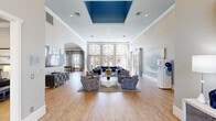 This generously proportioned single-floor community space makes a strong first impression through a glass-paneled entrance foyer with towering transom windows that flood the entryway with natural light. Just off the entry, a private office nook features a quilted navy accent wall, a functional desk, and warm lighting — ideal for leasing or administrative use. The heart of the space is a grand main lounge characterized by soaring ceilings with a bold cobalt blue tray ceiling detail, a crystal chandelier, and light hardwood flooring throughout. Large arched windows and French doors line the walls, bathing the interior in abundant daylight while offering views of the surrounding greenery and pool area. Elegant blue velvet sectional sofas, patterned accent chairs, and geometric coffee tables anchor multiple seating arrangements, creating an inviting and cohesive aesthetic. A wide archway connects the main lounge to a secondary lounge area featuring a fireplace, ceiling fans, and additional seating. A dedicated game room, framed by floor-to-ceiling windows and sheer drapes, houses a shuffleboard table and casual seating, with direct visual access to an outdoor pool. A quieter sitting area with soft blue-grey walls, curated wall art, and refined furnishings rounds out the space, offering a more intimate retreat within this stylish, multi-functional facility.
