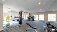 This fitness center offers a comprehensive workout environment designed to accommodate a wide range of exercise needs. The space is single-level and generously proportioned, with a layout that allows for comfortable movement between equipment stations. The flooring consists of durable, multi-toned carpet in shades of blue and green, providing cushioning underfoot throughout the workout area. Recessed lighting in the white ceiling ensures the space is well-lit, while large, multi-pane windows along the walls flood the room with abundant natural light, creating an open and energizing atmosphere. The ceiling features a vaulted section toward one end, adding a sense of height and openness to the space. Equipment includes multiple treadmills for cardiovascular training, cable pulley machines suited for a variety of strength exercises, a dedicated weight bench for resistance training, and additional strength-training apparatus. A full-length mirror is positioned along one wall, supporting proper form and technique during workouts. The neutral, light-toned walls contribute to the clean and focused ambiance. Overall, this fitness center provides a functional, well-lit, and thoughtfully arranged space suitable for both cardiovascular and strength training routines.