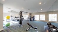 This fitness center offers a comprehensive workout environment designed to accommodate a wide range of exercise needs. The space is single-level and generously proportioned, with a layout that allows for comfortable movement between equipment stations. The flooring consists of durable, multi-toned carpet in shades of blue and green, providing cushioning underfoot throughout the workout area. Recessed lighting in the white ceiling ensures the space is well-lit, while large, multi-pane windows along the walls flood the room with abundant natural light, creating an open and energizing atmosphere. The ceiling features a vaulted section toward one end, adding a sense of height and openness to the space. Equipment includes multiple treadmills for cardiovascular training, cable pulley machines suited for a variety of strength exercises, a dedicated weight bench for resistance training, and additional strength-training apparatus. A full-length mirror is positioned along one wall, supporting proper form and technique during workouts. The neutral, light-toned walls contribute to the clean and focused ambiance. Overall, this fitness center provides a functional, well-lit, and thoughtfully arranged space suitable for both cardiovascular and strength training routines.