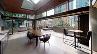 Located at 2125 6th Ave in Seattle's Belltown neighborhood, this sophisticated amenity lounge seamlessly blends indoor comfort with outdoor living. The space is defined by dramatic floor-to-ceiling glass walls and expansive sliding doors that open fully to an adjoining outdoor terrace, dissolving the boundary between interior and exterior. A striking warm-toned wood slat ceiling crowns the interior, complemented by a skylight that floods the space with natural light and a polished concrete floor that anchors the industrial-modern aesthetic. Exposed concrete walls add texture and an urban edge throughout.

The interior is centered around a substantial communal wood dining table surrounded by woven rope and upholstered seating, ideal for gatherings. A built-in wet bar along one wall features dark wood cabinetry, a white countertop, dual sinks, a beer tap, and a wall-mounted television, providing full entertaining capabilities.

The outdoor terrace extends the living area with a wood deck, lounge seating arranged around a fire pit, and a professional-grade outdoor grill station. Lush planters line the perimeter, while panoramic views of Seattle's dynamic skyline provide a compelling urban backdrop. The overall ambiance is refined yet approachable, purpose-built for communal socializing and relaxation.