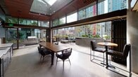 Located at 2125 6th Ave in Seattle's Belltown neighborhood, this sophisticated amenity lounge seamlessly blends indoor comfort with outdoor living. The space is defined by dramatic floor-to-ceiling glass walls and expansive sliding doors that open fully to an adjoining outdoor terrace, dissolving the boundary between interior and exterior. A striking warm-toned wood slat ceiling crowns the interior, complemented by a skylight that floods the space with natural light and a polished concrete floor that anchors the industrial-modern aesthetic. Exposed concrete walls add texture and an urban edge throughout.

The interior is centered around a substantial communal wood dining table surrounded by woven rope and upholstered seating, ideal for gatherings. A built-in wet bar along one wall features dark wood cabinetry, a white countertop, dual sinks, a beer tap, and a wall-mounted television, providing full entertaining capabilities.

The outdoor terrace extends the living area with a wood deck, lounge seating arranged around a fire pit, and a professional-grade outdoor grill station. Lush planters line the perimeter, while panoramic views of Seattle's dynamic skyline provide a compelling urban backdrop. The overall ambiance is refined yet approachable, purpose-built for communal socializing and relaxation.