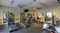This fitness center is a purpose-built exercise facility designed to accommodate a variety of cardiovascular and general workout needs. The space is outfitted with multiple treadmills, elliptical machines, and additional cardio equipment, all arranged to maximize usability and flow. Durable rubber flooring covers the workout area, providing both safety and comfort underfoot. Full-length mirrors line the walls, enhancing the sense of openness while allowing users to monitor their form during exercise. Track lighting runs along the ceiling, delivering bright, even illumination throughout the room, complemented by ceiling fans that help maintain comfortable airflow. A wall-mounted flat-screen television is positioned for visibility from the cardio equipment, adding an entertainment element to workout sessions. The walls feature a two-tone color scheme with a warm neutral upper half and a soft yellow-green lower accent, contributing to an energizing yet approachable atmosphere. Natural light filters in through a window, adding a welcoming brightness to the space. Overall, the fitness center strikes a balance between functionality and comfort, offering a well-maintained environment suited for daily exercise routines.