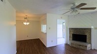 This 2-floor home offers a welcoming main living area defined by rich, dark hardwood-style flooring and neutral white walls that create a clean, versatile backdrop. A standout feature of the living room is the brick fireplace positioned in the corner, adding warmth and character to the space. A ceiling fan overhead provides practical climate control, while a decorative pendant light fixture illuminates the adjacent dining area, which benefits from a pass-through window connecting it to the kitchen — a convenient layout detail that enhances the open feel between rooms. The kitchen pass-through allows natural light to flow between spaces while maintaining a functional separation. A doorway leads to what appears to be an outdoor patio or enclosed space, offering additional natural light and outdoor access directly from the main living area. The textured ceilings are consistent throughout, adding a subtle design element overhead. The overall layout is practical and well-connected, with the ground floor designed to encourage easy movement between the living, dining, and kitchen areas, while the second floor provides additional living space above.