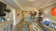 This single-floor residence showcases a thoughtfully designed open-concept layout that seamlessly connects the main living areas. Rich, dark hardwood flooring flows throughout the space, grounding the interior with warmth and continuity. The living area is generously proportioned, offering ample room for comfortable furnishings, and is anchored by a neutral color palette that creates a calm, welcoming ambiance. A chevron-patterned area rug and tasteful décor accents add visual interest to the space. The kitchen is a standout feature, boasting dark cabinetry, stainless steel appliances, pendant lighting over a central island with bar seating, and recessed lighting that highlights the work surfaces. The island serves as a natural gathering point and a practical transition between the kitchen and living space. Warm pendant lights cast an inviting glow over the kitchen area, complementing the overall earthy tones of the interior. Tray ceilings add architectural dimension to the space, lending a sense of height and refinement. A hallway visible from the main living area suggests additional private rooms beyond. The overall design strikes a balance between modern aesthetics and functional living, making this a well-appointed and versatile space.