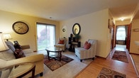 Located at 825 Crossing Court in Virginia Beach, Virginia, this single-floor home offers a comfortable and well-appointed living environment. The living room serves as the heart of the space, showcasing warm hardwood flooring that transitions seamlessly into a carpeted seating area, creating defined zones for relaxation and conversation. Large sliding glass doors flood the room with abundant natural light while framing a verdant outdoor view, lending the interior a bright and inviting atmosphere. The neutral, softly painted walls provide a versatile backdrop that complements the space's warm tones. A decorative accent wall features a stylish oval mirror and a sleek shelving unit, adding visual interest and a touch of elegance to the room. The open layout allows for a natural flow throughout the main living area, making the space feel expansive and welcoming. Overall, the home presents a harmonious blend of functional design and comfortable living, well-suited for those seeking a relaxed yet refined lifestyle in the Virginia Beach community.
