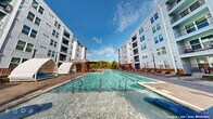 This outdoor pool amenity in Wake Forest, NC offers a well-appointed retreat designed for relaxation and recreation. The centerpiece is a generously sized swimming pool with crystal-clear water, accessible via stainless steel entry rails and a zero-entry wading area that gradually slopes into the pool. Flanking the pool deck are distinctive arch-shaped private cabanas outfitted with cushioned blue seating, providing shaded lounging spaces ideal for individual or small-group relaxation. Additional sunbathing chairs and chaise lounges are arranged along the perimeter, offering ample space for residents to unwind. A covered pergola structure at one end of the pool deck adds a shaded gathering point, complemented by lush mature trees that frame the open sky above. The surrounding hardscape features a combination of stamped concrete and paver stone surfaces, creating a refined, cohesive aesthetic. Raised brick planters with ornamental grasses and shrubs add natural texture and visual interest to the space. The pool area is enclosed by a sleek metal fence with a gated entry, ensuring privacy and safety. Towering multi-story residential buildings with modern grey siding and brick accents frame the courtyard on multiple sides, creating an intimate yet open-air atmosphere throughout this thoughtfully designed amenity space.