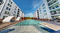 This outdoor pool amenity in Wake Forest, NC offers a well-appointed retreat designed for relaxation and recreation. The centerpiece is a generously sized swimming pool with crystal-clear water, accessible via stainless steel entry rails and a zero-entry wading area that gradually slopes into the pool. Flanking the pool deck are distinctive arch-shaped private cabanas outfitted with cushioned blue seating, providing shaded lounging spaces ideal for individual or small-group relaxation. Additional sunbathing chairs and chaise lounges are arranged along the perimeter, offering ample space for residents to unwind. A covered pergola structure at one end of the pool deck adds a shaded gathering point, complemented by lush mature trees that frame the open sky above. The surrounding hardscape features a combination of stamped concrete and paver stone surfaces, creating a refined, cohesive aesthetic. Raised brick planters with ornamental grasses and shrubs add natural texture and visual interest to the space. The pool area is enclosed by a sleek metal fence with a gated entry, ensuring privacy and safety. Towering multi-story residential buildings with modern grey siding and brick accents frame the courtyard on multiple sides, creating an intimate yet open-air atmosphere throughout this thoughtfully designed amenity space.