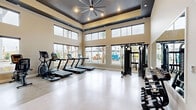 Located at 3225 W Colonial Dr in Orlando, FL, this well-appointed fitness center offers a comprehensive workout environment within a generously sized, open-plan space. The room is defined by its striking tray ceiling finished in a dark tone, fitted with recessed lighting and a large ceiling fan that together create a polished, functional atmosphere. Floor-to-ceiling windows and clerestory windows along multiple walls flood the interior with natural light while offering views of the surrounding outdoor landscape, including palm trees and adjacent buildings. The polished concrete-style flooring is both durable and easy to maintain, lending a clean, modern aesthetic throughout. Cardio equipment including multiple treadmills and elliptical trainers is positioned along the windowed walls, allowing users to exercise while enjoying the outdoor views. A multi-station cable machine anchors the strength-training area, accompanied by a full rack of dumbbells and a wall-mounted mirror that enhances the sense of space. An open stretch and functional training zone features a bold tropical-print accent wall and a medicine ball storage rack. A dedicated restroom is accessible directly from the fitness floor, adding convenience for users.