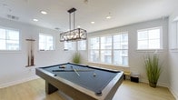The Diamond amenity space is a thoughtfully designed single-floor facility that blends comfort and entertainment seamlessly. The interior is characterized by bright white walls, light hardwood-style flooring, and an abundance of recessed lighting that creates a clean, welcoming atmosphere throughout.

The lounge area is well-appointed with plush gray seating, teal accent chairs, and a glass coffee table arranged on a patterned area rug. A large flat-screen television is mounted on the wall, complemented by a decorative console table with carefully curated accessories. Colorful artwork adds visual interest to the walls, while a pub-style high table with bar stools provides an additional social gathering spot. Natural light filters in through multiple windows fitted with light-filtering shades.

A separate billiards area features a full-size pool table as its centerpiece, dramatically highlighted by a geometric industrial-style pendant light fixture. Large windows along one wall invite generous natural light and offer views of the surrounding building exterior. The space maintains the same light flooring and neutral color palette, ensuring a cohesive design aesthetic throughout the entire facility.