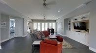 This single-floor space showcases an open-concept layout that seamlessly connects the living, dining, and kitchen areas into one cohesive environment. The interior is finished with cool grey walls and dark concrete flooring, creating a contemporary aesthetic throughout. The living area is furnished with a large sectional sofa and vibrant red-orange accent chairs arranged around a central coffee table, adding a bold contrast to the neutral palette. Two ceiling fans with industrial-style fixtures are mounted overhead, complementing the recessed lighting embedded throughout the ceiling. Large French doors along the rear wall flood the space with natural light and provide views of the surrounding outdoor area. Adjacent to the living space, a dining area with a dark table and chairs offers a defined yet connected gathering spot. A built-in shelving unit and mounted flat-screen television anchor one wall, adding functional storage and entertainment options. Toward the back, a kitchen with stainless steel appliances and a bar-style counter is visible, rounding out this well-appointed, multi-functional space.