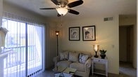 This single-story residence at Brickinridge Court offers a welcoming and thoughtfully designed living space. The living room serves as the heart of the home, featuring a ceiling fan with a built-in light fixture that provides both airflow and ambient illumination. Soft, neutral-toned walls create a warm and inviting atmosphere, complemented by carpeted flooring that adds comfort underfoot. A large sliding glass door fitted with vertical blinds allows natural light to fill the room while offering direct access to an outdoor area, providing a seamless connection between indoor and outdoor living. The space is furnished with a plush sofa accented by decorative pillows, a mirrored coffee table, and a coordinating side table topped with an ornate lamp and greenery. Framed wall art depicting elegant figures adds a refined, decorative touch to the room. A tall floor lamp in the corner contributes additional warm lighting, enhancing the cozy ambiance. The overall aesthetic is casual yet polished, making it a comfortable and functional space suited for both relaxing and entertaining.