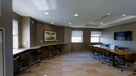 Located at 205 Church Street in the heart of New Haven, CT, this business center offers a well-appointed, single-floor workspace designed to support a range of professional needs. The interior is finished in a contemporary style, with taupe-toned walls, neutral carpeting in a subtle tile pattern, and a clean white ceiling outfitted with recessed lighting that ensures consistent, even illumination throughout the space.

Two distinct work zones define the layout. Along one wall, a built-in counter workstation provides individual seating for multiple users, each position equipped with a modern rolling chair — ideal for focused, independent work. On the opposite side of the room, a larger table configuration accommodates group collaboration or meetings, with additional ergonomic seating arranged for team use.

Natural light enters through multiple windows fitted with horizontal blinds, offering adjustable brightness and a connection to the outside environment. A framed piece of artwork adorns the wall near the individual workstations, adding a touch of sophistication to the professional setting. The overall ambiance is calm, functional, and conducive to focused work, making this business center a practical and refined resource for residents and professionals alike.