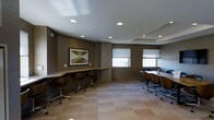 Located at 205 Church Street in the heart of New Haven, CT, this business center offers a well-appointed, single-floor workspace designed to support a range of professional needs. The interior is finished in a contemporary style, with taupe-toned walls, neutral carpeting in a subtle tile pattern, and a clean white ceiling outfitted with recessed lighting that ensures consistent, even illumination throughout the space.

Two distinct work zones define the layout. Along one wall, a built-in counter workstation provides individual seating for multiple users, each position equipped with a modern rolling chair — ideal for focused, independent work. On the opposite side of the room, a larger table configuration accommodates group collaboration or meetings, with additional ergonomic seating arranged for team use.

Natural light enters through multiple windows fitted with horizontal blinds, offering adjustable brightness and a connection to the outside environment. A framed piece of artwork adorns the wall near the individual workstations, adding a touch of sophistication to the professional setting. The overall ambiance is calm, functional, and conducive to focused work, making this business center a practical and refined resource for residents and professionals alike.