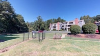 This residential community is nestled within a lush, tree-lined setting that provides a sense of privacy and natural tranquility. The multi-story buildings feature a classic architectural style highlighted by warm brick chimneys, neutral-toned siding, and private balconies that offer residents elevated outdoor living space. The grounds are thoughtfully landscaped, with mature deciduous and pine trees casting dappled shade across mulched beds, manicured shrubbery, and expansive open lawns. A dedicated green space anchors the community's common area, complemented by a decorative bench and a fenced enclosure that adds a functional outdoor amenity. A community pool is visible among the trees, providing a recreational focal point for residents. Well-maintained paved roadways wind through the property, lined with mature oak trees whose canopies arch gracefully overhead, creating a shaded corridor throughout the community. Ample surface parking is conveniently distributed across the grounds. The overall ambiance is one of a peaceful, established neighborhood where natural surroundings and residential comfort coexist seamlessly, offering residents a serene retreat within a cohesive and carefully maintained outdoor environment.