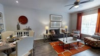 This single-story space showcases a well-appointed living area that blends contemporary and transitional design elements. The living room is anchored by rich, wood-look hardwood flooring that runs throughout, providing a warm and cohesive foundation for the space. A bold orange geometric area rug adds a striking pop of color beneath a white rectangular coffee table, creating a defined seating zone. The dark charcoal sofa is flanked by matching geometric side tables, each topped with table lamps that contribute warm ambient lighting to the room. A patterned accent chair adds visual interest alongside the sofa, while neutral gray walls provide a clean backdrop. Two framed geometric art pieces are displayed in a stacked arrangement on the main wall, complementing the room's modern aesthetic. A ceiling fan with an integrated light fixture is centrally mounted overhead, offering both functionality and a finished look. A buffet or sideboard along one wall adds storage and display space, while burnt orange curtains frame a window, tying the color palette together cohesively. The overall ambiance is stylish, warm, and inviting.