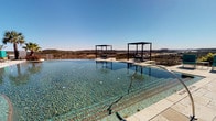 This outdoor pool area is designed as a resort-style amenity space, offering two distinct pool settings within the same complex. The first features a striking infinity-edge pool with clear turquoise water that seamlessly blends into sweeping panoramic views of the surrounding landscape and open sky. Two sleek, dark-framed pergola cabanas are positioned poolside, providing shaded lounging areas that enhance the upscale aesthetic. The pool deck is finished in light-toned stone tile, lending a polished, high-end feel to the space.

A separate pool area presents a generously sized rectangular pool with an adjacent spa, surrounded by an expansive concrete sun deck lined with lounge chairs and market umbrellas. This area is framed by mature trees offering natural shade, with the multi-story building serving as an architectural backdrop that reinforces the communal character of the space.

Adjacent to the pool is a covered outdoor pavilion featuring natural stone columns and a warm wood-paneled ceiling, creating an inviting shaded retreat. The travertine-paved patio accommodates additional seating and lounge chairs, ideal for outdoor gatherings. Access to the pool area is provided through a dedicated single-story pool house structure with glass-paneled doors, offering direct sightlines to the pool from within.