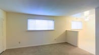 This single-floor space offers a clean, functional layout with a simple and understated aesthetic. The interior is finished with neutral, light-toned walls that create a bright and open atmosphere throughout. Soft, wall-to-wall carpeting runs underfoot, adding warmth and comfort to the living areas.

Natural light is a key feature of this space, with horizontal windows fitted with white blinds that allow for adjustable brightness and privacy. The windows are positioned to draw in ambient daylight, keeping the interior feeling fresh and airy. A partial wall or countertop partition is visible in one area, suggesting a defined transition between living zones or an open-plan layout element that adds architectural interest.

The smooth, curved ceiling detail adds a subtle design touch that distinguishes the space from a purely standard build. The overall ambiance is calm and neutral, offering a versatile blank canvas well-suited to a variety of furnishing styles and personal preferences. The space presents itself as a tidy, move-in ready environment with practical finishes throughout.