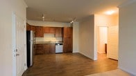 This single-floor residence showcases a thoughtfully designed open-concept layout, seamlessly connecting the kitchen and living areas to create a spacious, functional environment. The kitchen is a standout feature, equipped with rich, medium-toned wood cabinetry that extends to the ceiling, offering generous storage throughout. Granite countertops provide a durable and attractive work surface, complemented by stainless steel appliances, including a refrigerator and dishwasher. A track lighting system runs across the ceiling, ensuring the kitchen is well-lit and practical for daily use. The flooring throughout the main living space consists of warm, dark hardwood-style planks that add depth and visual continuity to the open floor plan. Neutral-toned walls enhance the brightness of the space, creating a clean and welcoming ambiance. A wide doorway at the far end of the living area leads to additional rooms, suggesting a well-connected and easy-to-navigate layout. White baseboards provide a crisp finish, tying together the overall aesthetic. The combination of warm wood tones and neutral walls gives the space a balanced, inviting character suited for everyday living.