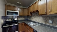 This single-floor residence offers a thoughtfully arranged layout connecting a functional kitchen to open living and sleeping areas. The kitchen serves as a standout feature, showcasing warm medium-toned wood cabinetry with ample upper and lower storage, dark granite-style countertops, and a neutral square-tile backsplash. Stainless steel appliances include an electric range, over-the-range microwave, and dishwasher, complemented by a stainless steel sink. Wood-look flooring runs throughout the kitchen, adding warmth to the space.

The living area and bedrooms are finished with wall-to-wall neutral carpeting and painted in a soft gray tone, creating a calm and versatile backdrop. Horizontal windows allow natural light to filter in, brightening the carpeted rooms. The entry area transitions from hardwood-style flooring into the carpeted spaces, providing clear delineation between zones.

The bathroom includes a granite-style countertop surface consistent with the kitchen's finishes, along with a wall-mounted towel bar. Throughout the unit, white baseboards and neutral wall tones maintain a cohesive, clean aesthetic suited to a variety of furnishing styles.