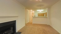 This single-floor residence offers a functional and cohesive layout designed for comfortable living. The living area serves as the central gathering space, finished with wall-to-wall carpeting in a warm beige tone that adds softness underfoot. A classic white-mantled fireplace with a black surround anchors one wall, serving as a natural focal point and adding a touch of traditional character to the room. Crown molding runs along the ceiling perimeter, lending a refined architectural detail throughout the space.

The living area flows seamlessly toward the kitchen, which is separated by a raised breakfast bar — ideal for casual dining or entertaining. The kitchen features medium-toned wooden cabinetry providing ample storage, a white appliance suite including a refrigerator and microwave, and overhead lighting that keeps the space bright and practical. A pendant light fixture above the breakfast bar adds a decorative element while enhancing task lighting.

A hallway connects the living and kitchen areas to the remaining rooms of the residence, suggesting a thoughtful layout with clear separation between communal and private spaces. The overall palette of soft whites and warm neutrals creates a calm, inviting ambiance throughout.