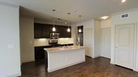 Located at 6040 Parker Blvd in North Richland Hills, Texas, this single-story space showcases a contemporary design with a thoughtfully appointed open-concept layout. The kitchen serves as a central highlight, featuring rich dark espresso cabinetry that extends to the ceiling, offering generous storage throughout. A large central island with light granite countertops and an integrated double sink provides both workspace and a natural gathering point. Stainless steel appliances, including a gas range, built-in microwave, and French door refrigerator, complement the kitchen's modern aesthetic. A textured tile backsplash adds depth and visual interest behind the cooking area. Three sleek pendant lights hang above the island, while a track lighting system illuminates the main cooking zone, ensuring the space is well-lit throughout. Dark hardwood-style flooring flows continuously through the kitchen and into the adjoining living area, creating a cohesive and grounded atmosphere. The clean white walls and crisp trim contrast elegantly with the darker cabinetry and flooring, lending the space a balanced, polished look. The open floor plan promotes a seamless flow between the kitchen and living areas, making the space feel welcoming and functional.
