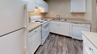 This single-floor residence offers a well-organized layout designed for comfortable everyday living. The kitchen is efficiently arranged in an L-shape, featuring white cabinetry, light countertops, a full suite of white appliances including a range, dishwasher, and refrigerator, along with a double-basin sink and wood-look flooring that adds warmth to the space. A central island or breakfast bar provides additional prep and dining space, seamlessly connecting to the adjacent living area, which is finished with plush carpeting and natural light from a large window. The bedrooms are carpeted and generously sized. One bedroom stands out with a dramatic vaulted ceiling and a signature arched window above standard blinds, creating an airy, light-filled atmosphere with views of mature greenery outside. The two bathrooms are clean and functional. One features a white vanity with ample cabinet storage, a large mirror, and wood-look tile flooring. The second includes a full soaking tub with a shower surround, a matching vanity, and a mirrored cabinet, also finished with wood-look tile. Throughout the home, neutral tones, white trim, and consistent flooring choices create a cohesive and easy-to-style aesthetic suitable for a variety of living arrangements.